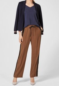 Navy v-ringad topp under en svart blazer, kombinerat med bruna byxor med svarta sidostråk, och ljusa skor.