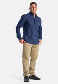 Marineblauw overhemd met lange mouwen, knoopsluiting en borstzak, gecombineerd met beige broek en zwarte sneakers, tegen een witte achtergrond.