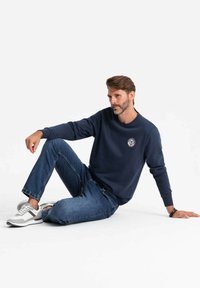 Sudadera navy con cuello redondo y un parche de logo, combinada con jeans azules y zapatillas grises. Diseño casual, material de mezcla de algodón.