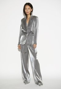 Zilveren metallic blazer en bijpassende broek met een strik taille, gekenmerkt door een glanzende textuur en wijde pijpen; versierd met unieke gevouwen zilveren schoenen.