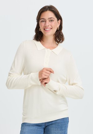 Jeune femme aux cheveux bruns portant un pull crème à manches longues avec col et un jean bleu, souriante, les mains jointes devant.