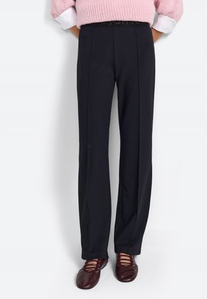 Pantalon noir sur mesure avec une coupe droite, présentant des plis à l'avant, une texture lisse et une taille étroite. Porté avec des chaussures bordeaux.