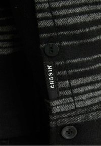 Tissu en laine rayé noir et gris avec des boutons noirs et une étiquette en tissu noir portant l'inscription « CHASIN' » cousue verticalement.