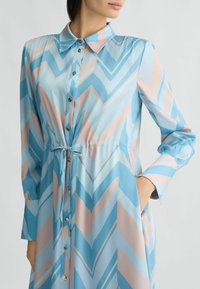 Abito camicia azzurro chiaro con motivo a zigzag in diverse tonalità di blu e rosa, con vita da annodare e maniche lunghe. Dettagli con bottoni argentati.