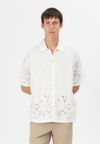 Hvid kortærmet skjorte med knap-op design, der har blomsterudskæringer i kanten og tekstureret stof.