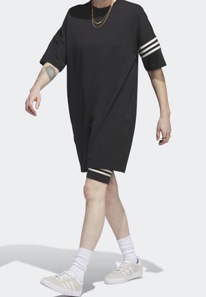 Persoon die loopt en een zwart oversized jurk draagt met witte strepen op de mouwen en korte broek, witte sokken, beige sneakers en gouden kettingen.