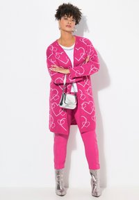 Růžový kardigan s bílými vzory srdce, nošený přes bílou košili a růžové kalhoty, doplněný stříbrnými kotníkovými botami a malou stříbrnou kabelkou.