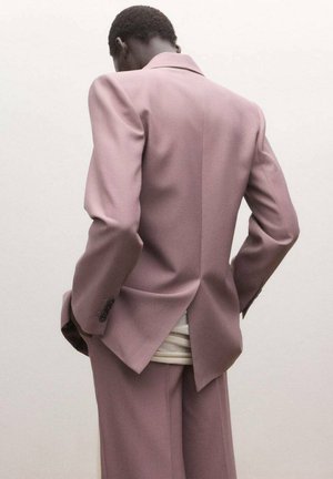 Hombre visto desde atrás con chaqueta de traje malva y pantalones a juego, con las manos en los bolsillos, sobre un fondo claro y sencillo.