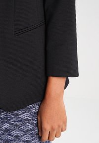 Nahaufnahme eines Arms einer Person, die ein langärmeliges, schwarzes, strukturiertes Oberteil und eine blau gemusterte Hose trägt, vor einem schlichten Hintergrund.