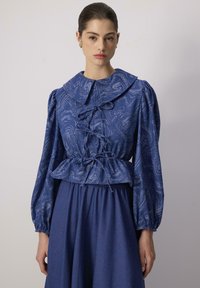 Blusa blu a fantasia con un ampio colletto, maniche a sbuffo e vita con cordoncino. La gonna è di un blu uniforme con una consistenza morbida.