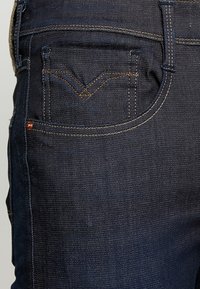 Tmavě modré džíny s jemným vyblednutím, zdobené oranžovým prošitím, kapsou se specifickým tvarováním ve tvaru V a bronzovým nýtem.