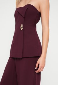 Top bordeaux sem alças com colarinho pontiagudo, apresentando um detalhe dourado e uma silhueta ajustada. Tecido liso com design ajustado.