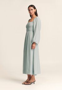 Dlhé, svetlomodré šaty s nariaseným živôtikom a dlhými nafúknutými rukávmi. Materiál je textúrovaný a modelka má na nohách čierne sandále na podpätku.