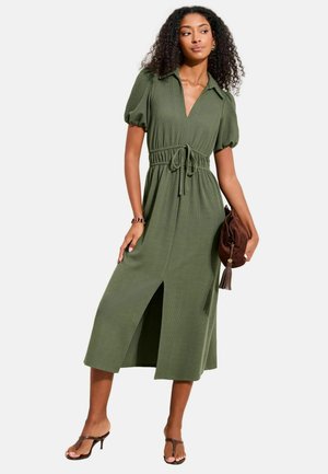 Frau trägt ein olivgrünes Midikleid mit Puffärmeln, einem vorderen Schlitz und einem Taillenzug, hält eine braune Handtasche mit Quasten.