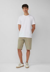 Weißes Baumwoll-T-Shirt, kombiniert mit beigen Denim-Shorts und cremefarbenen Sneakers mit strukturierten Akzenten. Das Model steht mit den Händen hinter dem Rücken.