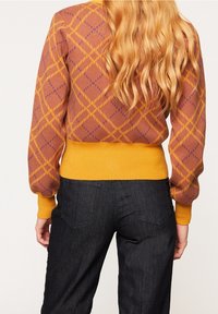 Kvinna med långt, vågigt blont hår som bär en brun och gul mönstrad tröja och mörka jeans, sedd bakifrån.