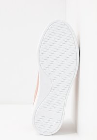 Semelle de basket blanche avec un motif antidérapant texturé, montrée à plat contre un mur d'angle blanc.