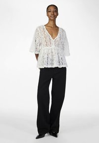 White floral lace top with a V-neck and short sleeves, featuring a peplum design, worn with black wide-leg trousers and pointed heels.