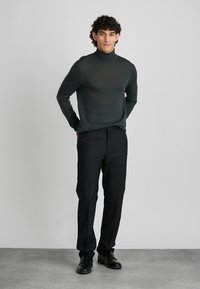 Noor päris noormees, kellel on lokkis juuksed, kannab tumerohelist kõrge kraega sviitrit, musti kohandatud pükse ja musti pidulaudade kingi, seisab käed selja taga.