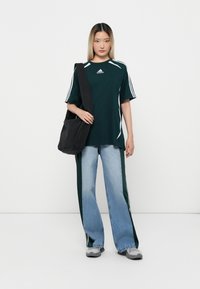 Dark green athletic shirt with white stripes, loose blue jeans with side stripes, and a black shoulder bag. Sneakers complete the outfit.