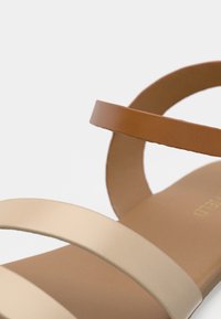 Κοντινό πλάνο ενός ταμπα σανδάλιου με δύο μπεζ λουριά πάνω από το πόδι και ένα ταμπα λουρί στον αστράγαλο, σε λευκό φόντο.