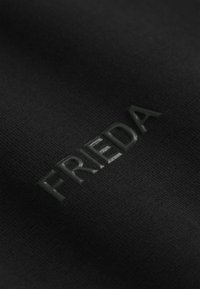 Czarna tkanina o teksturowanej powierzchni, z podniesionymi, ciemnymi literami wyraźnie tworzącymi napis "FRIEDA".