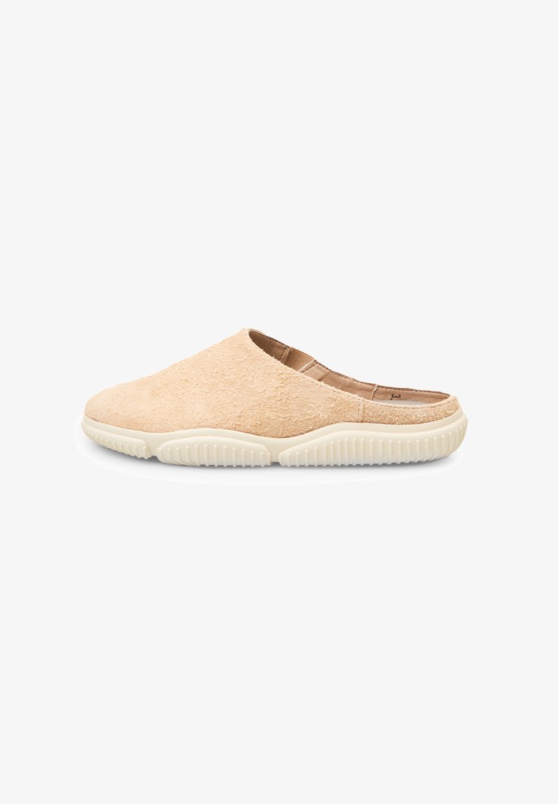 Beige slip-on shoes with a soft, textured upper and white rubber sole featuring a ridged pattern for grip. Rounded toe and backless design.