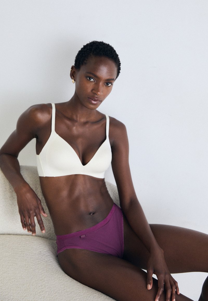 Femme aux cheveux courts et bouclés, portant un soutien-gorge blanc et un sous-vêtement violet, assise sur une chaise en tissu beige contre un mur clair et uni.