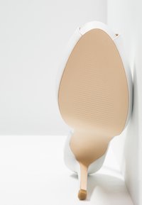 Chaussure blanche à talon haut avec une tige synthétique lisse, un design à bout effilé et une semelle texturée beige clair.