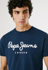 Junger Mann mit lockigem Haar, der ein marineblaues T-Shirt mit weißem "Pepe Jeans London"-Logo trägt und leicht nach links auf einen schlichten Hintergrund schaut.