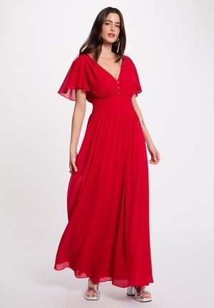 Donna in piedi con un abito lungo rosso fluente con maniche a svolazzo, scollo a V, dettagli con bottoni e tacchi argento aperti sulle punte, su sfondo bianco.