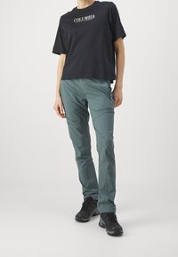 Svart bomullströja med "COLUMBIA" tryckt i vitt; teal utomhusbyxor med fickor och en avsmalnad passform; svarta vandringsskor.