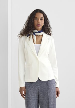 Žena kučeravých vlasov oblečená v bielom saku, vzorovaných sivých nohaviciach a tmavomodro-bielom šále na krk stojaca pri jednoduchej stene.