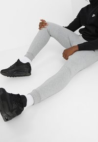 Person som bär grå mjukisar, vita strumpor, svarta Nike-sneakers och en svart Nike-jacka, sittandes med benen utsträckta på en vit golv.