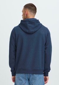 Uomo con i capelli corti, che indossa una felpa con cappuccio blu navy e jeans azzurri, in piedi di spalle contro uno sfondo chiaro e semplice.