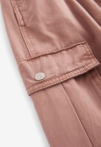 Šviesiai rožinė medžiaga su lygiu paviršiumi, su mygtuko detalėmis ir raukšlėta dizainu ant rankogalių. Artimas vaizdas.