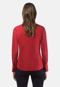 Chemise rouge à manches longues avec une coupe ajustée, texture lisse et poignets boutonnés. Présente un subtil détail de couture le long du dos.