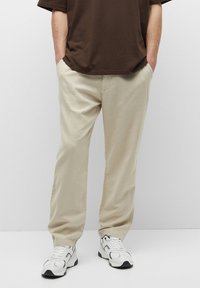 Man wearing beige casual pants and white sneakers, standing with hands in pockets against plain background.