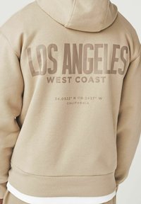 Sudadera con capucha beige que presenta el gran texto "LOS ÁNGELES" y "COSTA OESTE" en la parte posterior, con coordenadas geográficas y "CALIFORNIA" debajo.