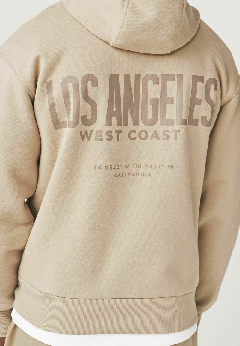 Sudadera con capucha beige que presenta el gran texto "LOS ÁNGELES" y "COSTA OESTE" en la parte posterior, con coordenadas geográficas y "CALIFORNIA" debajo.
