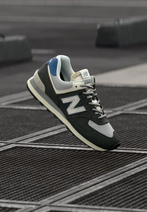 Grå og sort New Balance 574 sneaker med blå accenter, der svæver over en metalrist udendørs.