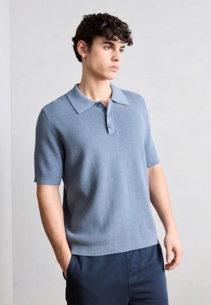 Unge mand med krøllet hår iført en lyseblå tekstureret kortærmet polo og marineblå bukser, stående med den ene hånd i lommen.