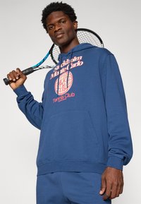 Kék kapucnis pulóverben, amelyen a "Stockholm Monte Carlo Tennis Club" felirat látható, férfi tenisztátot tart a válla fölé egy sima háttér előtt.
