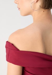 Rochie burgundy cu umerii descoperiți, cu o textură netedă. Linie a umerilor rotundă și un detaliu ușor pliat la decolteu. Cercei rotunzi din argint vizibili.
