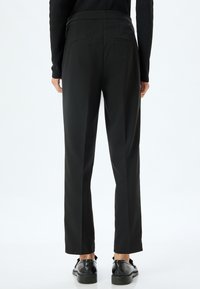 Zwarte rechte broek met een gladde, matte afwerking en achterzakken. Draagt zwarte schoenen, van achteraf weergegeven.