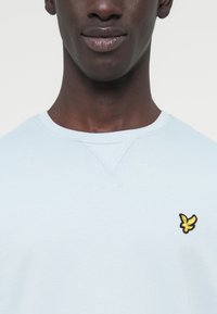 Sweat-shirt bleu clair avec un col rond et des poignets côtelés, orné d'un petit logo brodé noir et jaune sur la poitrine gauche.