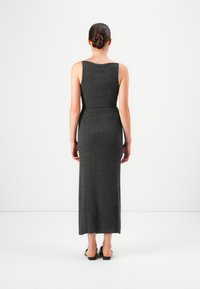 Ženska stoji s hrbtom proti kameri, oblečena v brezrokavno, teksturirano temno sivo maksi obleko in črne natikače, lasje speti v bun. 