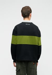 Asmuo, vilkintis juodai-žalią dryžuotą ilgų rankovių marškinį su "Santa Cruz" užrašu ant nugaros ir tamsiai žaliais velveto kelnėmis, stovi atsisukęs į kitą pusę.