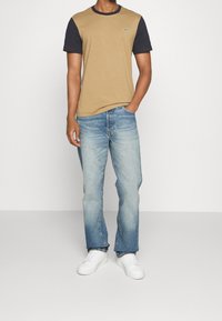 Tan short-sleeve T-shirt with dark blue sleeves, round neckline, and a small green logo, paired with light blue denim jeans and white sneakers.