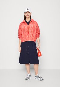 Cappotto a vento con motivo rosso e polsini elasticizzati, vestito blu con bottoni, cappellino bianco, borsa rossa e sneakers con dettagli argentati.